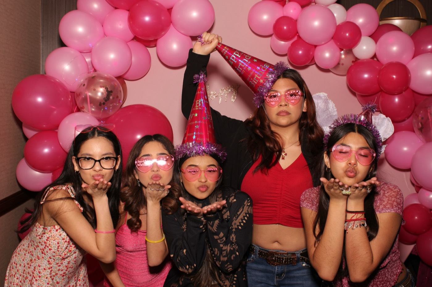 Friends blowing kisses in party hats in front of a pink balloon arch at a birthday party