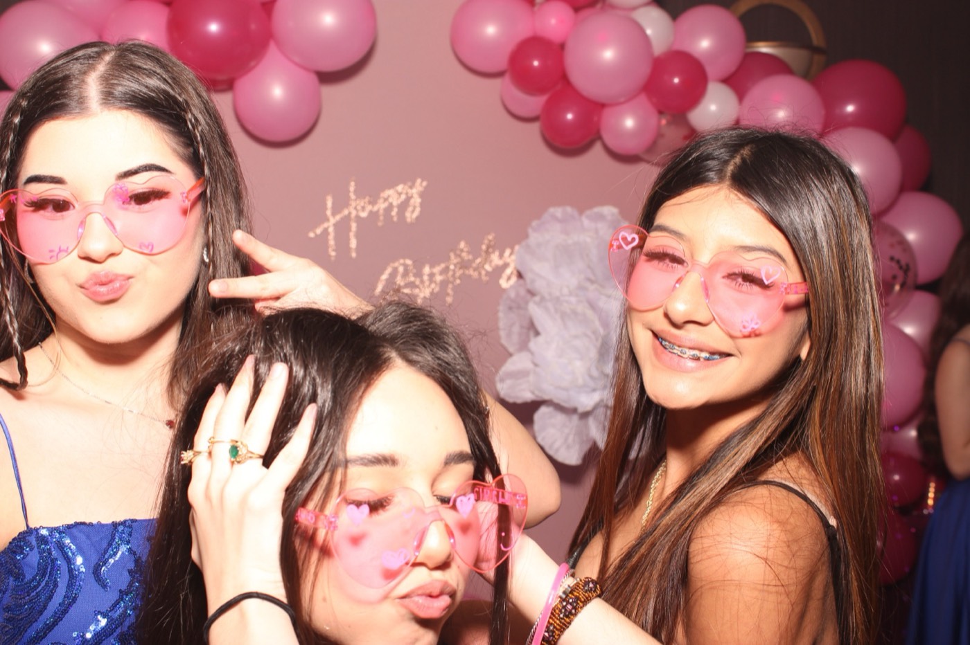 Friends in heart-shaped sunglasses in front of a pink balloon arch at a birthday party