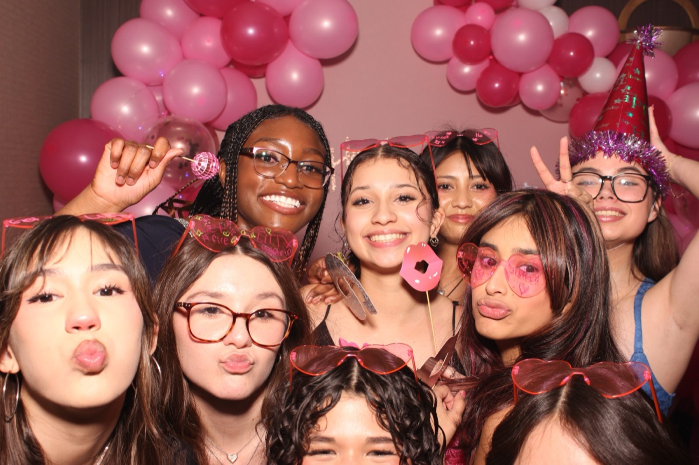 Friends crowding in with props in front of a pink balloon arch at a birthday party