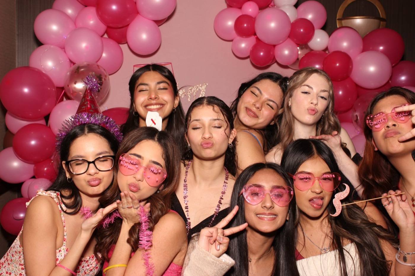 A big group of friends in heart glasses in front of a pink balloon arch at a birthday party