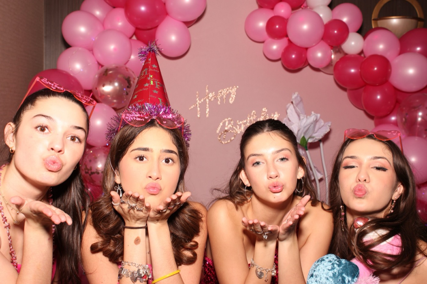 Four friends blowing kisses in party hats in front of a pink balloon arch at a birthday party
