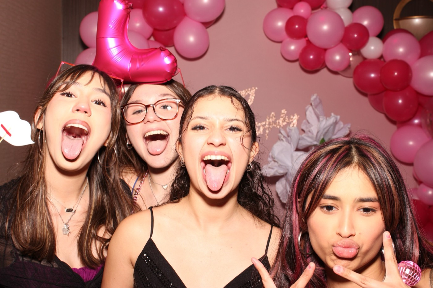 Friends sticking their tongues out in front of a pink balloon arch at a birthday party