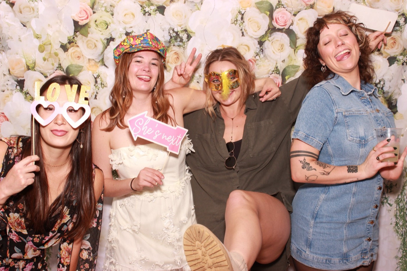 Friends laughing with heart glasses props in front of a white flower wall at a wedding