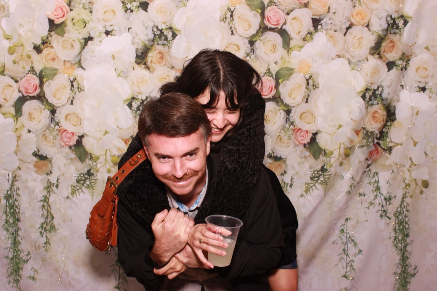 A guest giving his date a piggyback ride in front of a white flower wall at a wedding