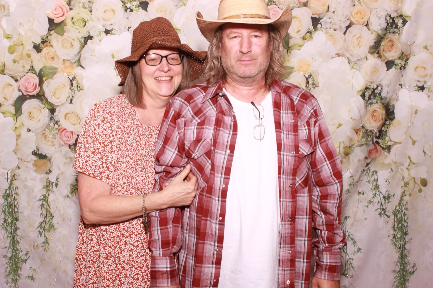 A couple in matching hats in front of a white flower wall at a wedding
