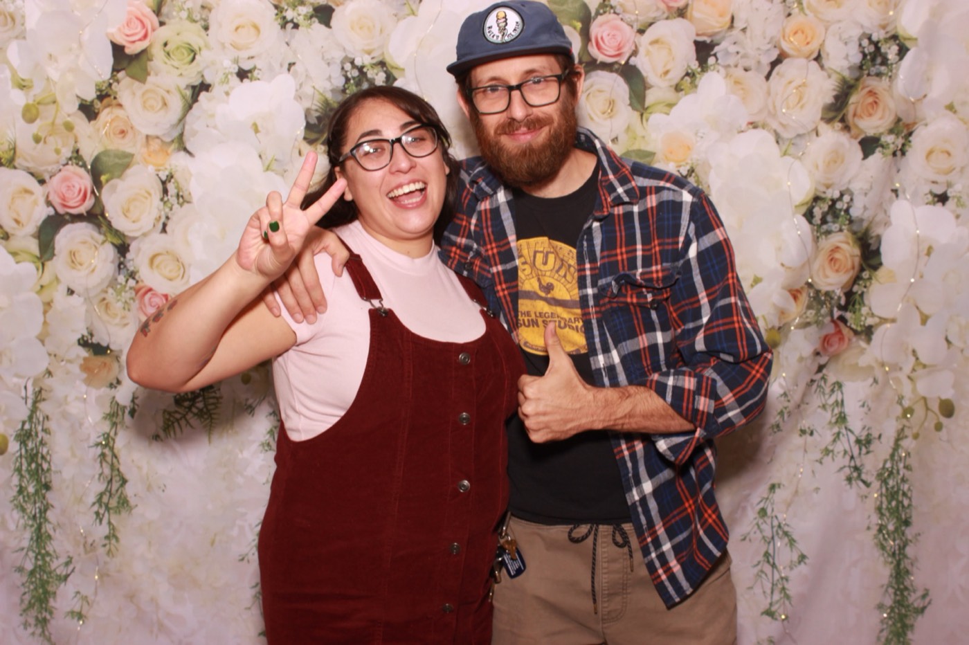 A couple throwing peace signs in front of a white flower wall at a wedding