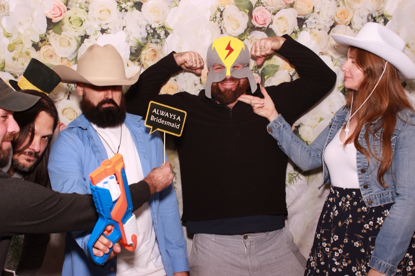 Groomsmen flexing in cowboy hats with props in front of a white flower wall at a wedding