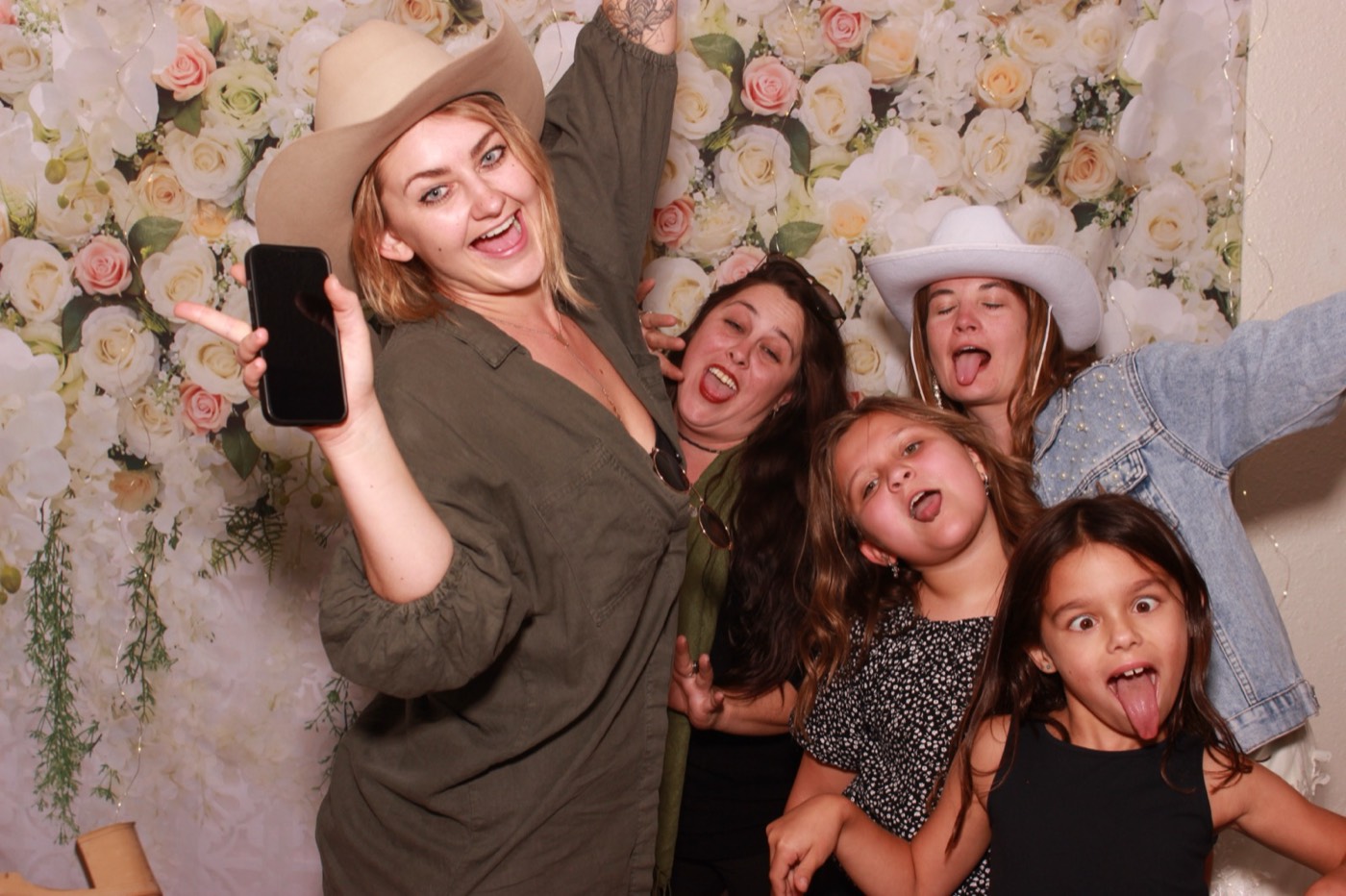 A guest with her phone and two kids in front of a white flower wall at a wedding