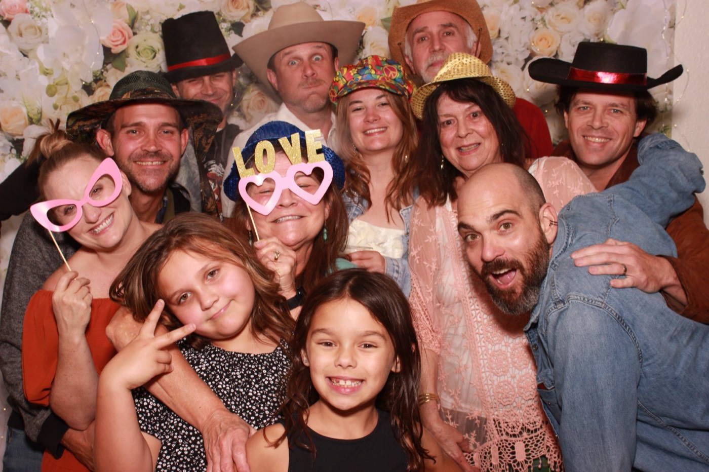 A family with heart glasses and a LOVE sign in front of a white flower wall at a wedding