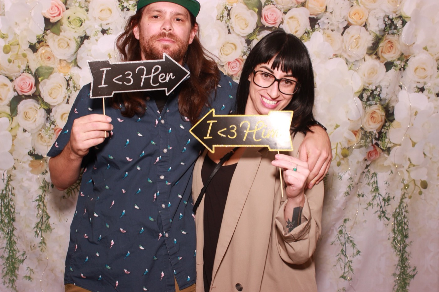 A couple holding I love her and I love him signs in front of a white flower wall at a wedding