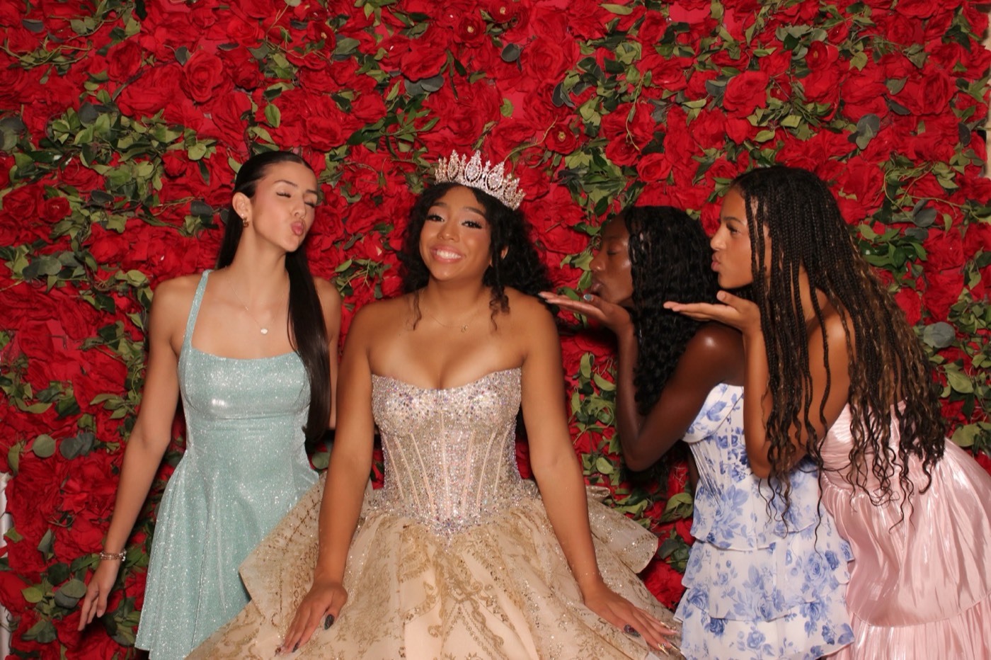 A quinceañera with friends in pastel dresses in front of a red rose wall at a San Antonio quince party