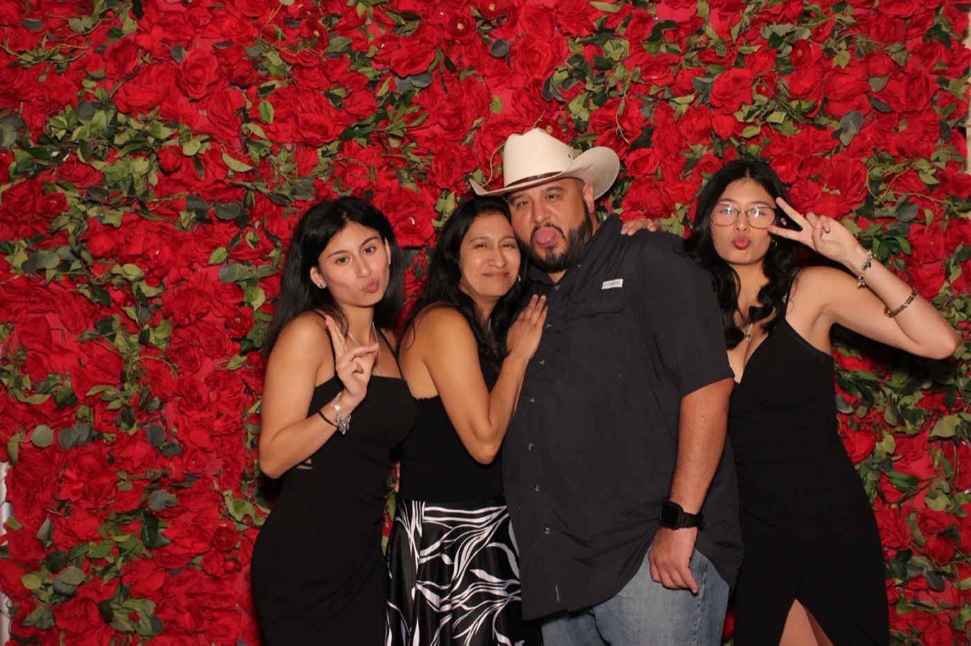 Friends posing with a guest in a cowboy hat in front of a red rose wall at a San Antonio quince party
