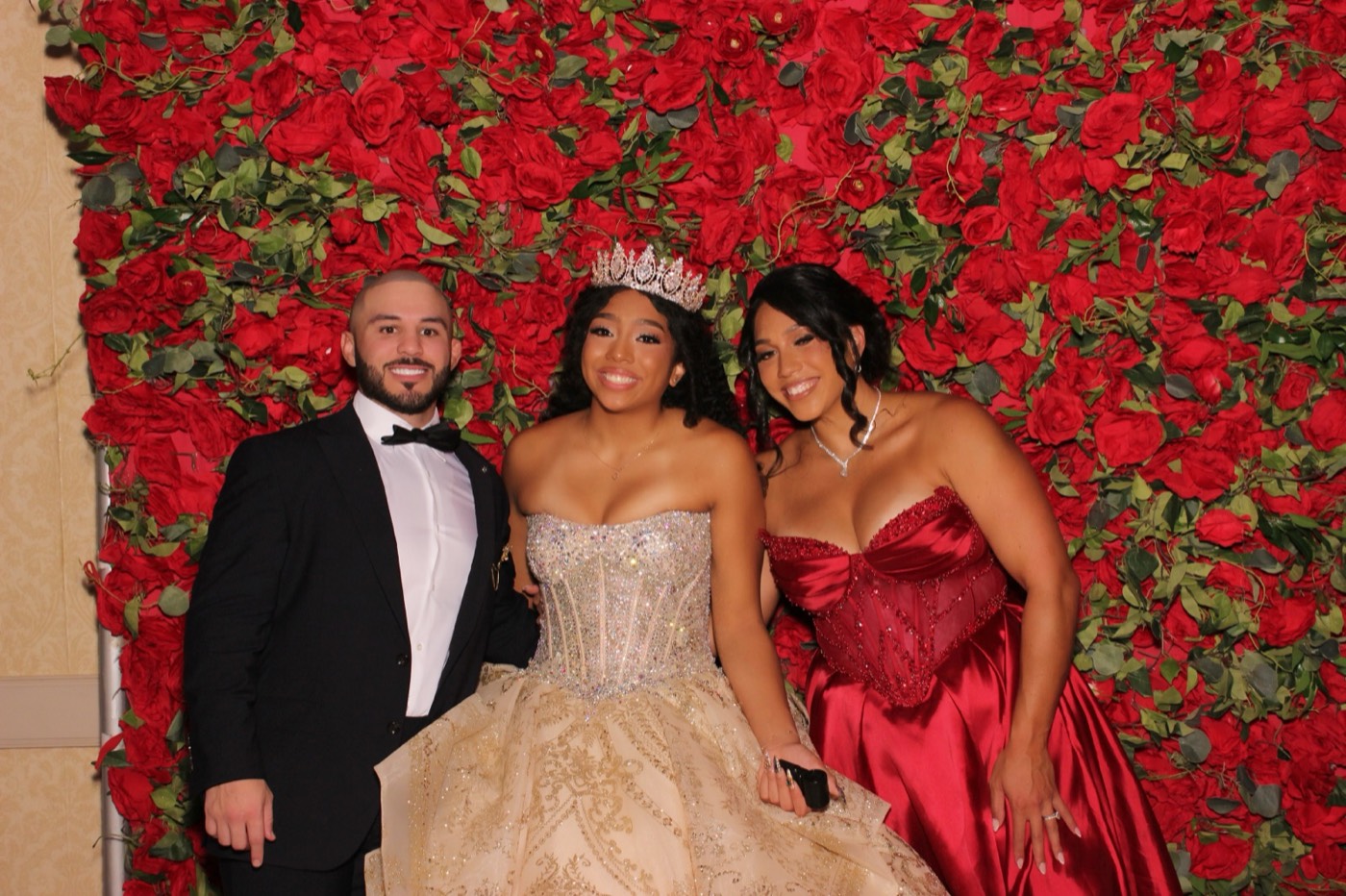 A quinceañera in a gold gown with two guests in front of a red rose wall at a San Antonio quince party