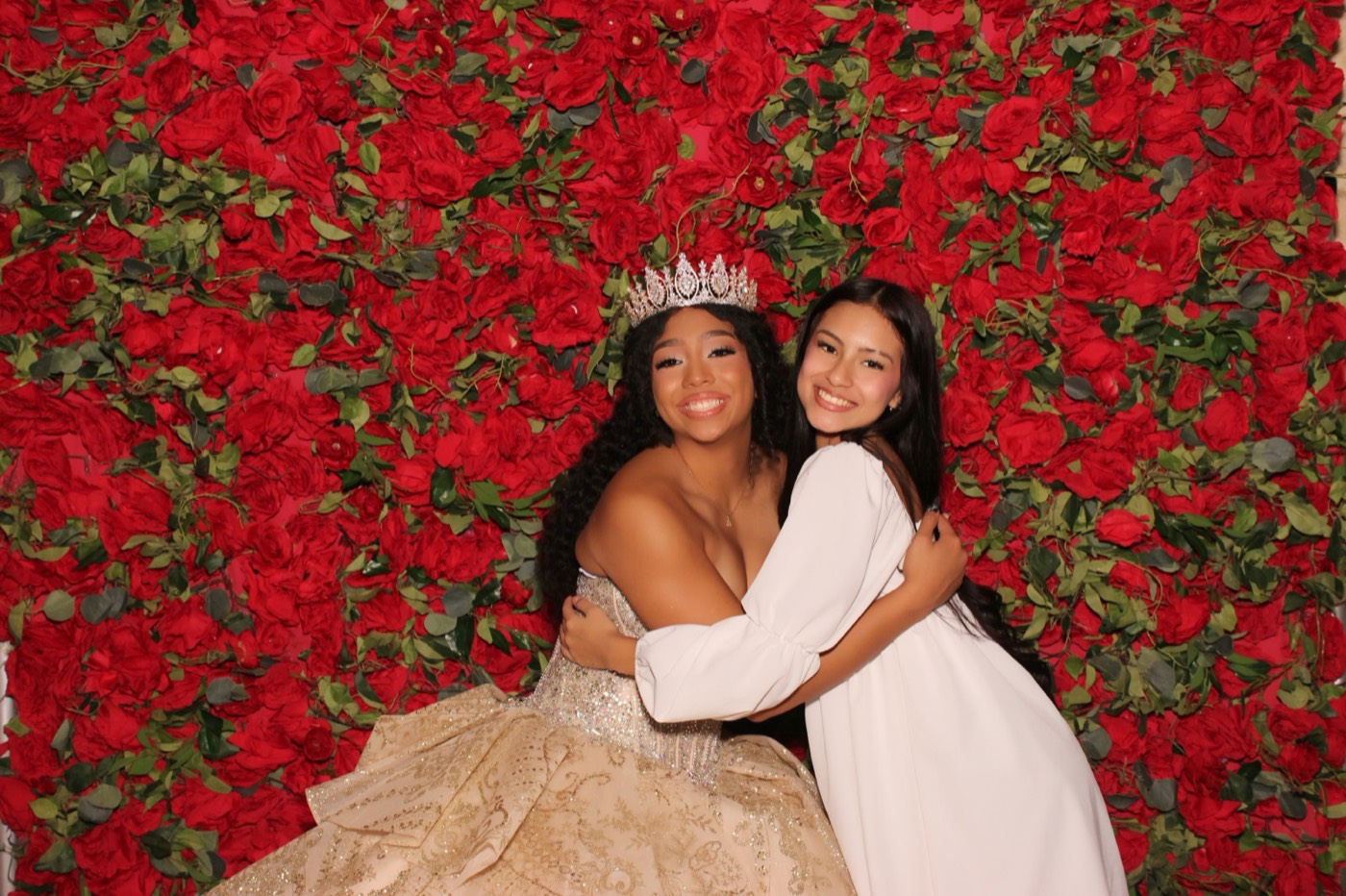 A quinceañera hugging a friend in front of a red rose wall at a San Antonio quince party