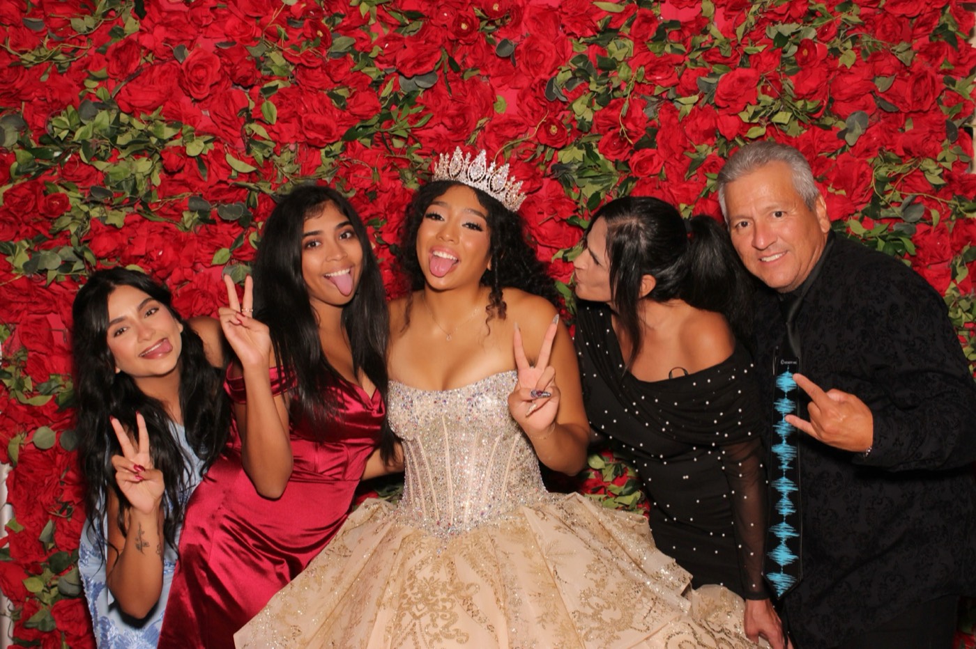A quinceañera with friends and family throwing peace signs in front of a red rose wall at a San Antonio quince party