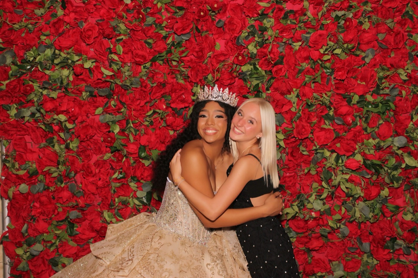 A quinceañera sharing a hug with a friend in front of a red rose wall at a San Antonio quince party