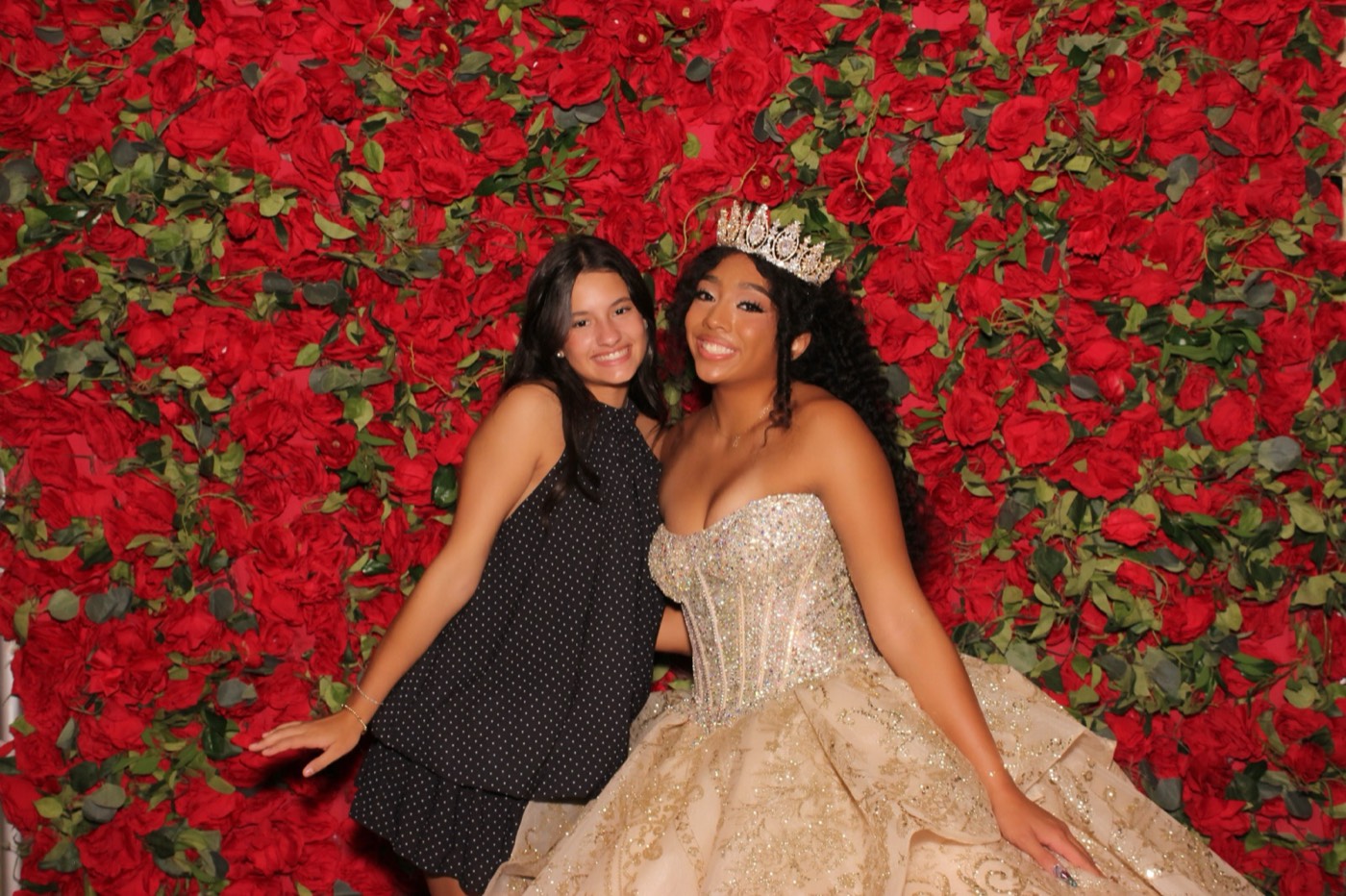 A quinceañera posing with a friend in front of a red rose wall at a San Antonio quince party
