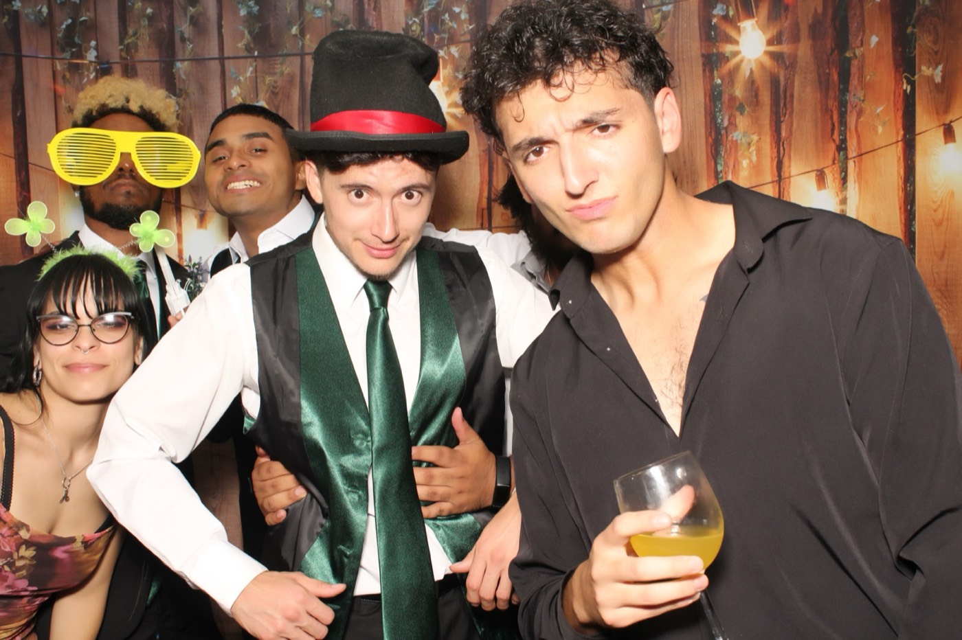 Groomsmen clowning around with props in front of a rustic wood wall with string lights at a wedding