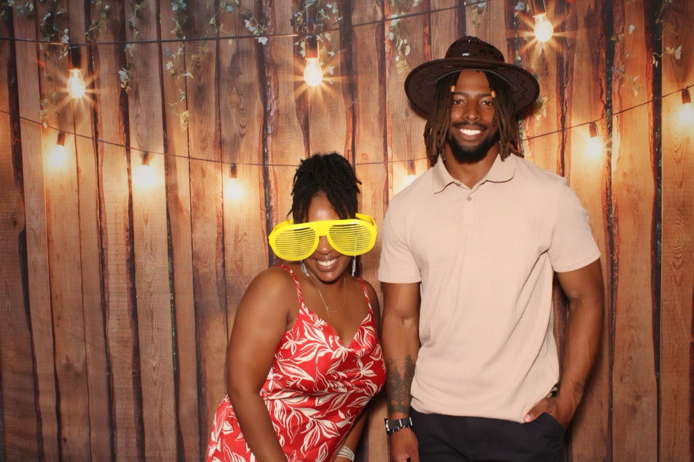 A couple in sunglasses and a cowboy hat in front of a rustic wood wall with string lights at a wedding