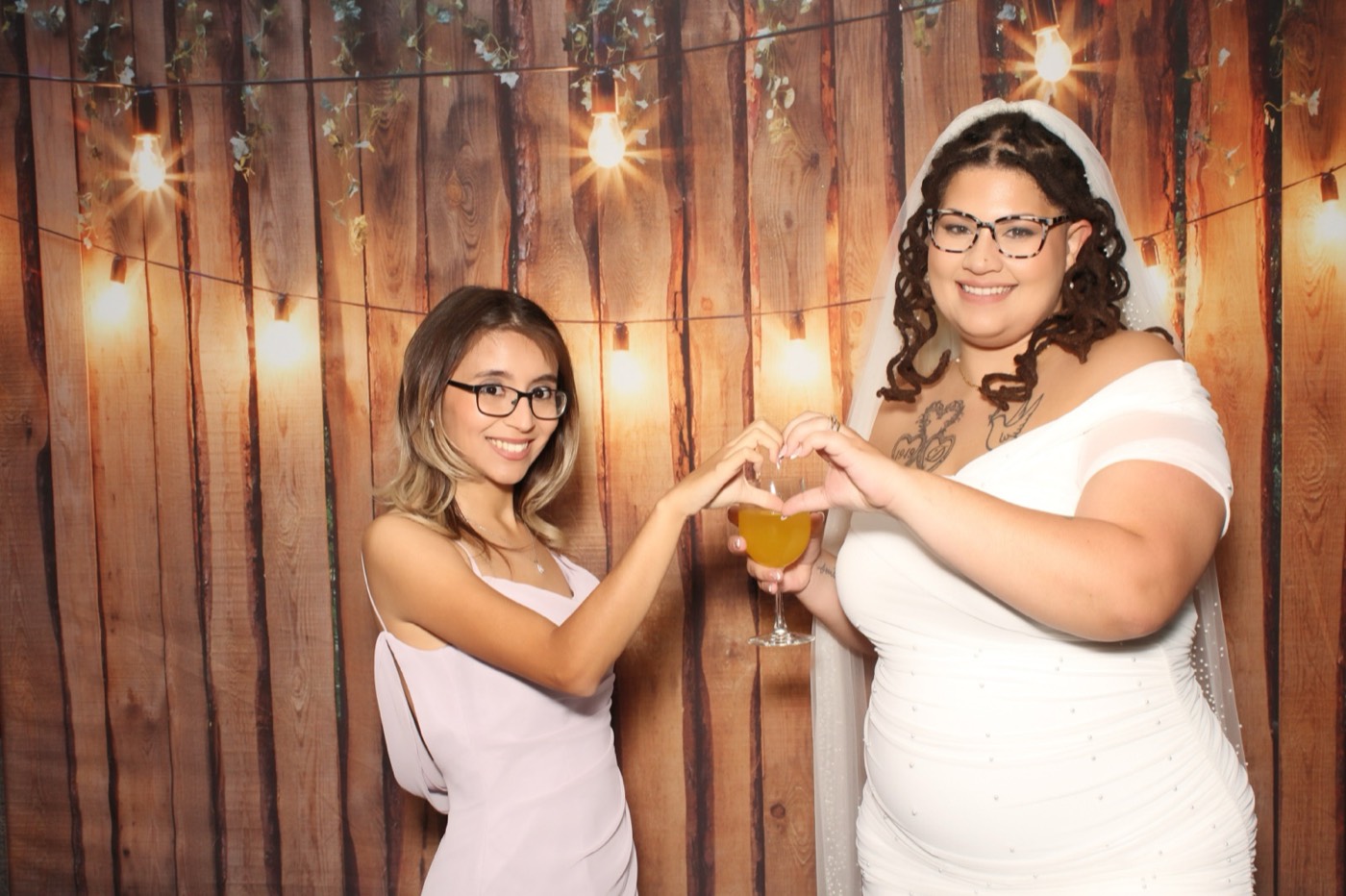 The bride and a friend making heart hands in front of a rustic wood wall with string lights at a wedding