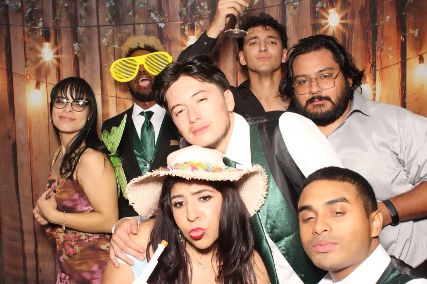 The wedding party in hats and party glasses in front of a rustic wood wall with string lights at a wedding