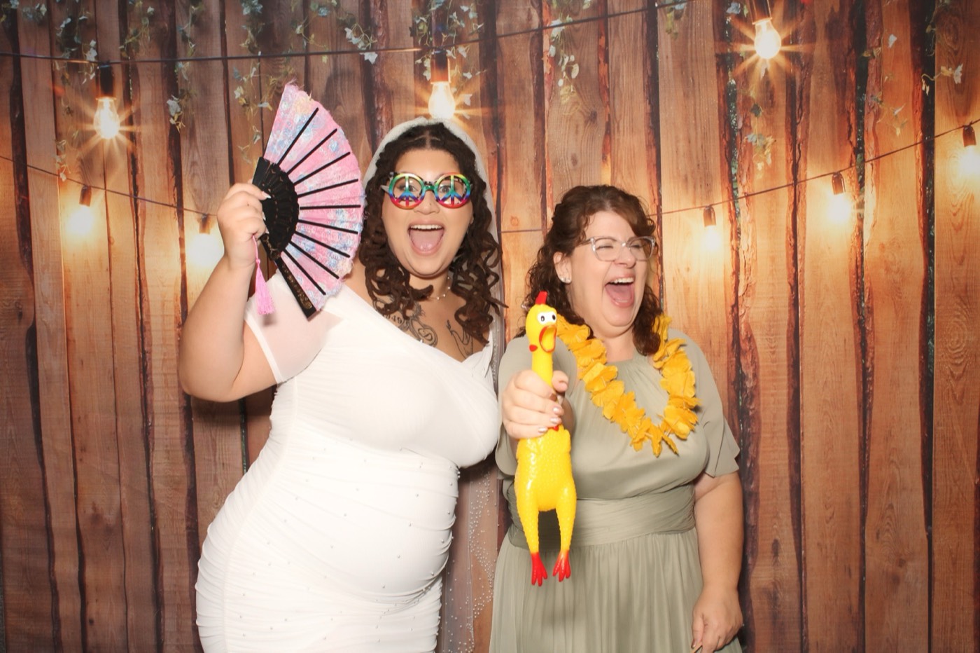 A guest with a fan and a flower lei in front of a rustic wood wall with string lights at a wedding