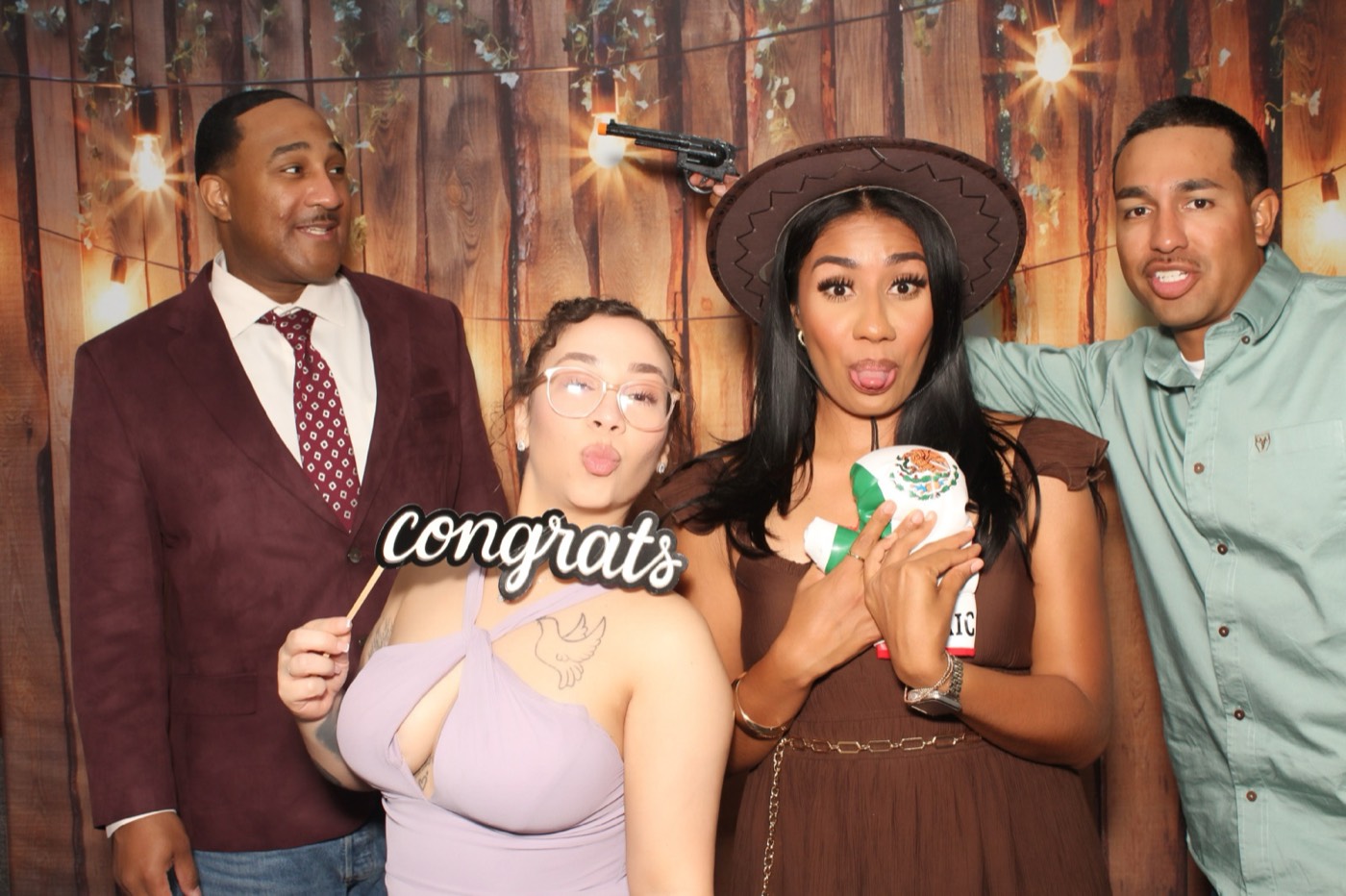 Friends holding a congrats sign in front of a rustic wood wall with string lights at a wedding