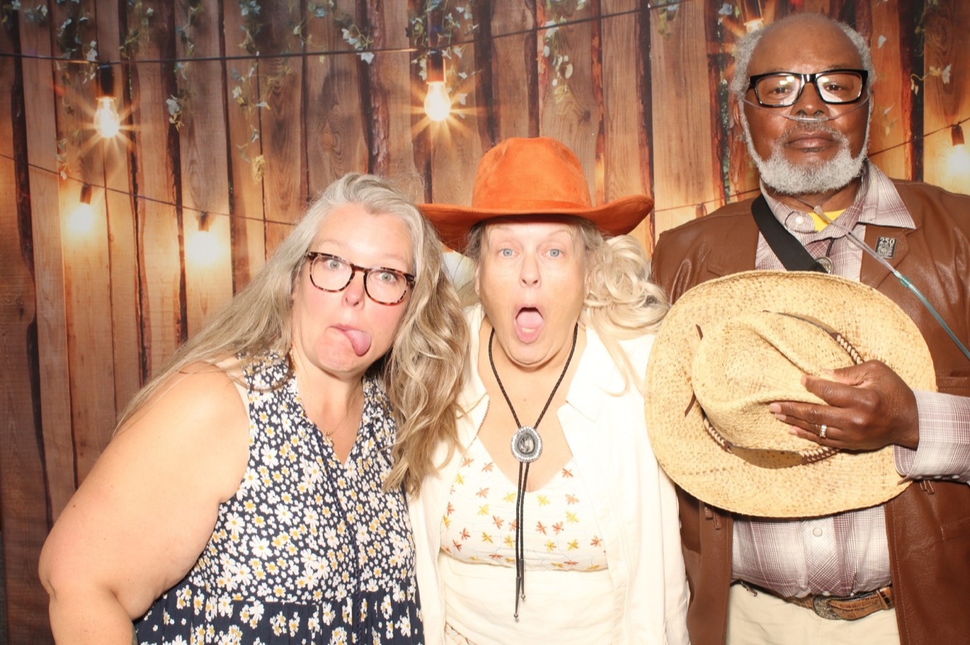Guests posing with a cowboy hat in front of a rustic wood wall with string lights at a wedding